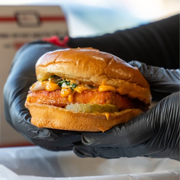 Gloved hands hold a Sumah Hot Chicken sandwich with pickles, sauce, and slaw on a brioche bun.
