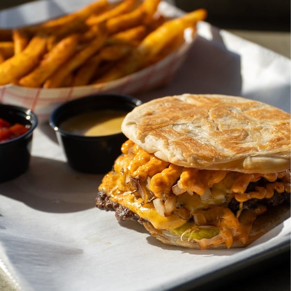 A Sumah Hot Chicken burger with cheese, onions, and pickles, served with fries and dipping sauces.
