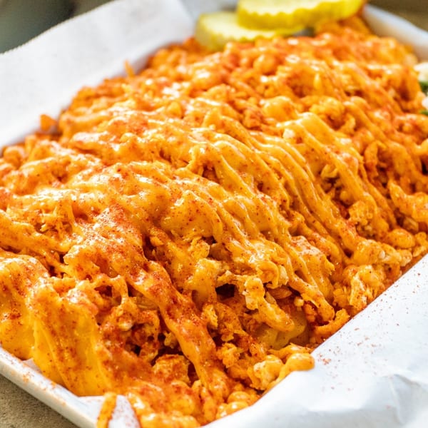 Close-up of Sumah Hot Chicken's waffle fries covered in melted cheese and paprika, with pickles in the background.
